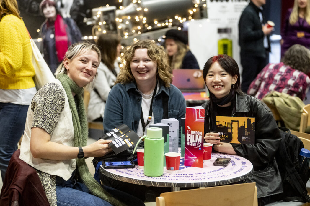 Three smiling people sit at a table looking at the camera. One holds up the BFI FAN CON schedule.