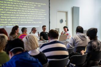 A group of people sit in chairs facing four panellists sitting at a table. One is passing a mike to another. Behind them is a large projector with yellow captions on a black background. Credit: Scottish Queer International Film Festival 2024, Ocean Teal.